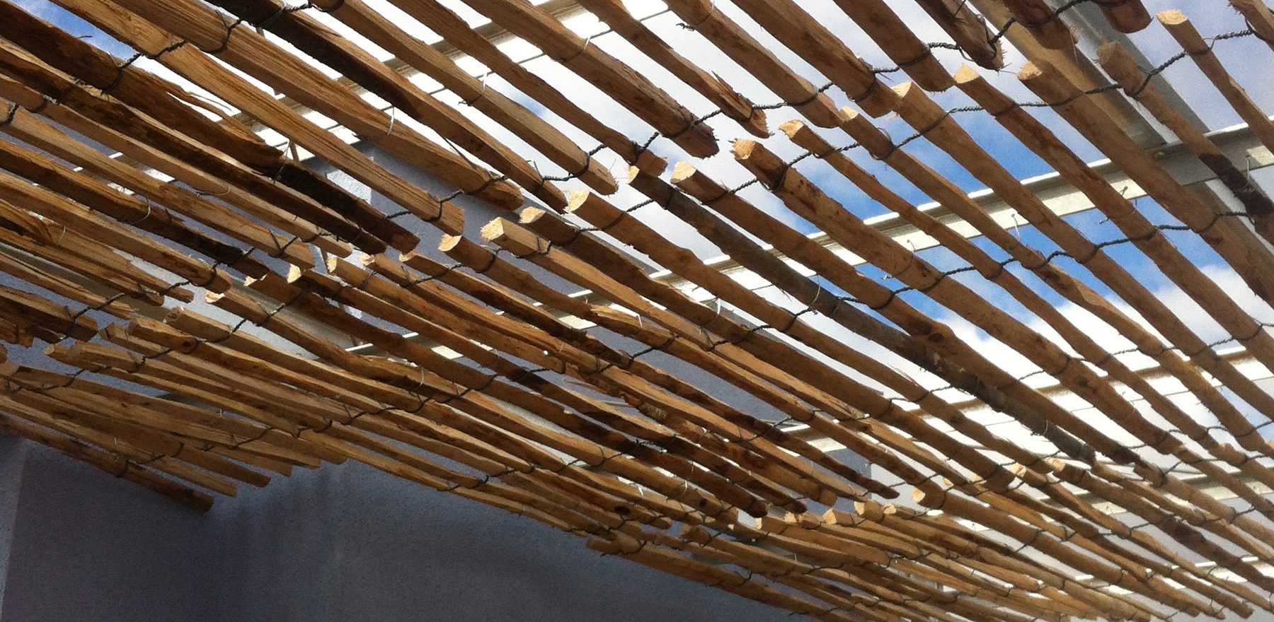 Blick von unten auf Holzdach aus locker gebundenen naturnahem, gespaltetem Holz vor blauem Himmel, das Schatten spendet